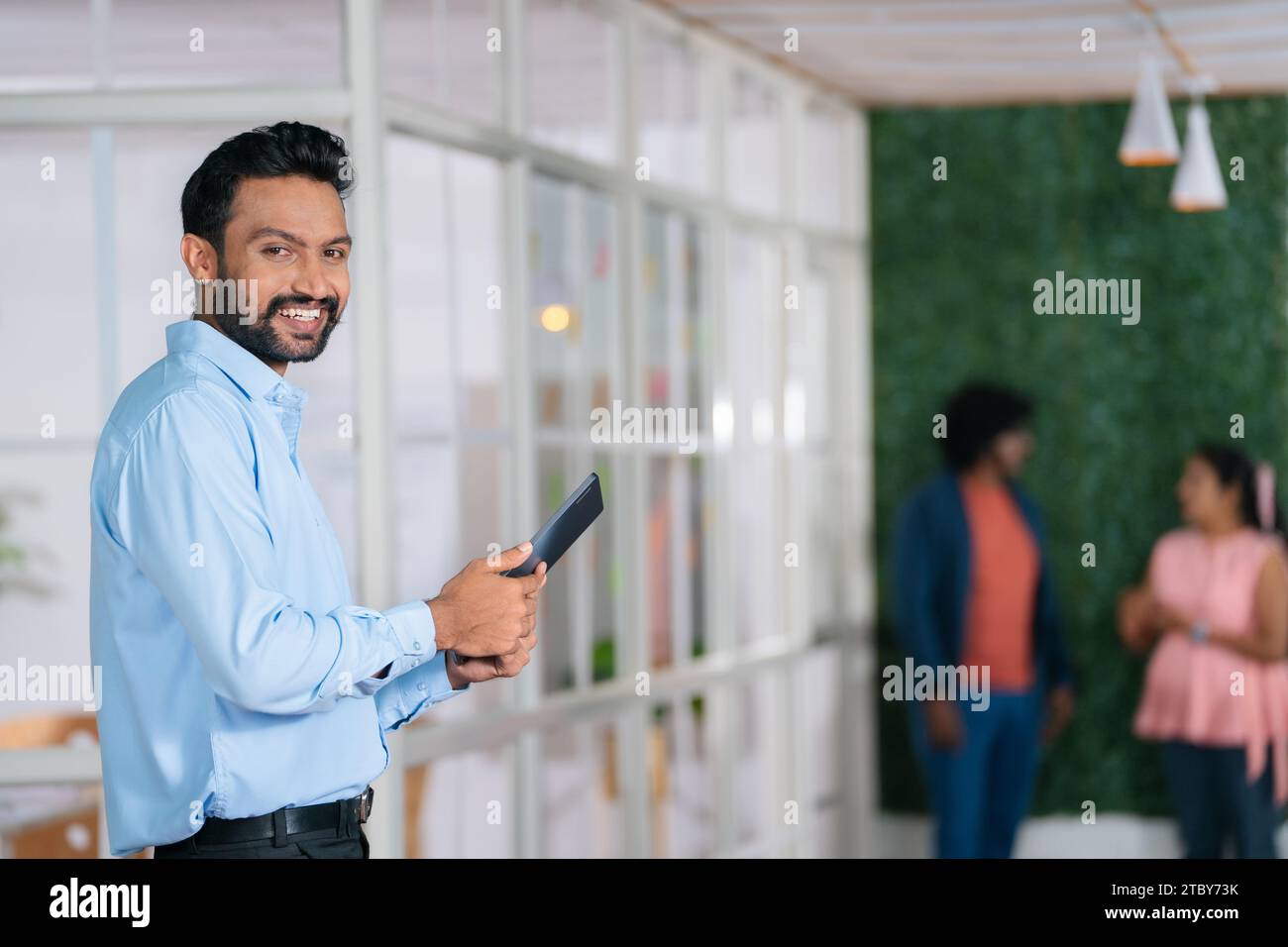 Happy young businessman confidently standing by looking camera with tab in hand at office - concept of startup entrepreneur, corporate employee and Stock Photo