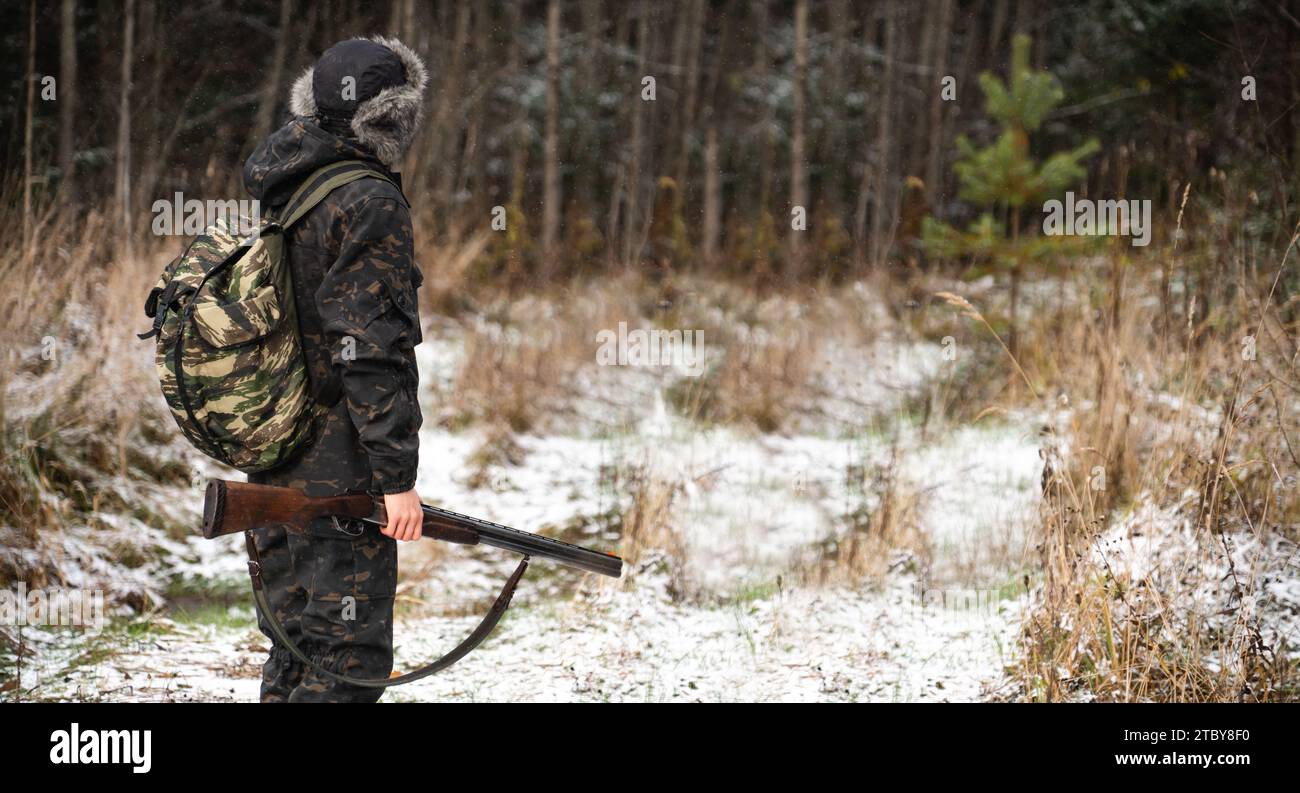 Male hunter in camouflage and with backpack, armed with a rifle, walks through the snowy winter forest.. Stock Photo