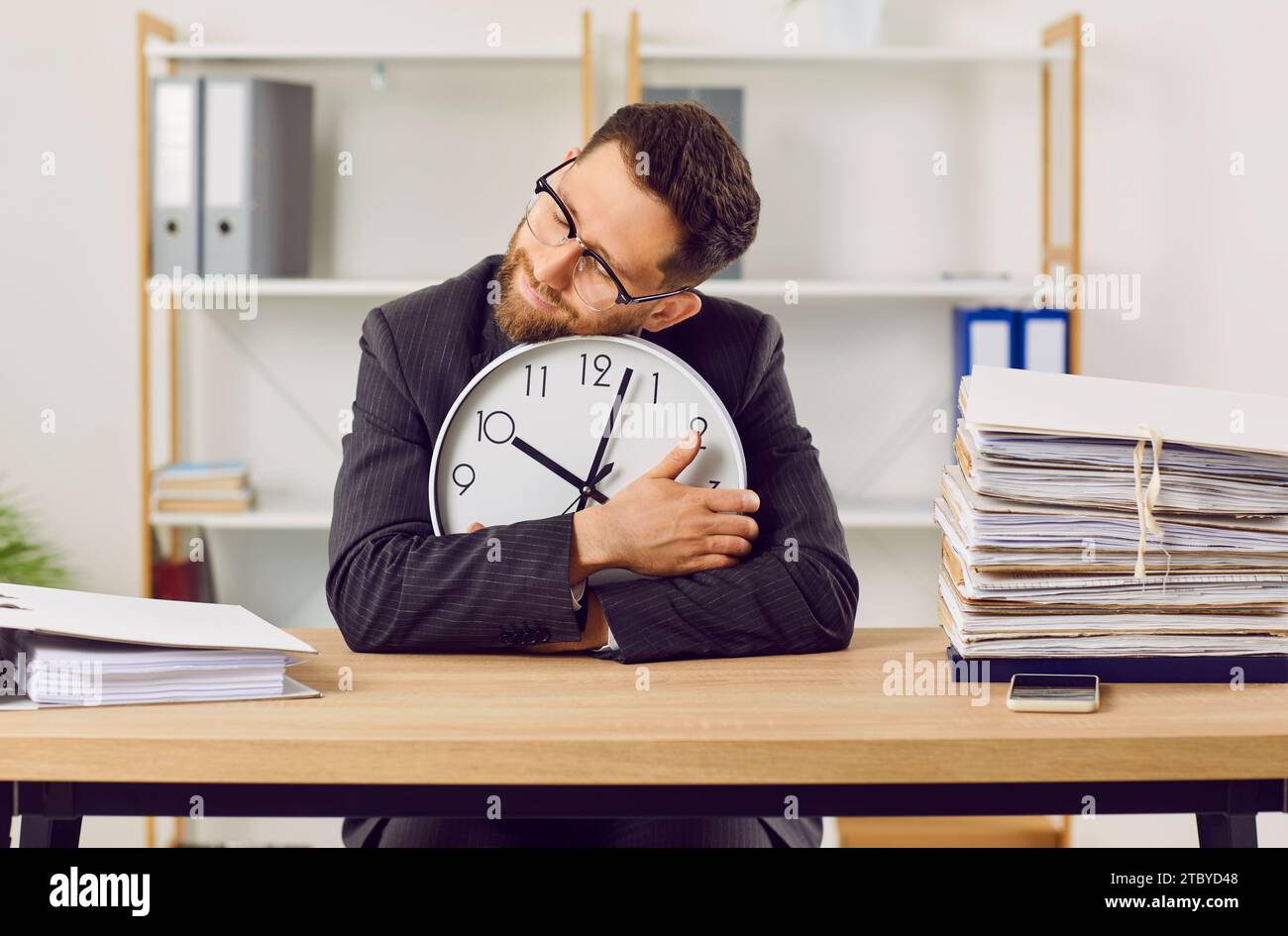 Funny, tired, overworked bookkeeper sitting at office desk, holding clock, and sleeping Stock Photo
