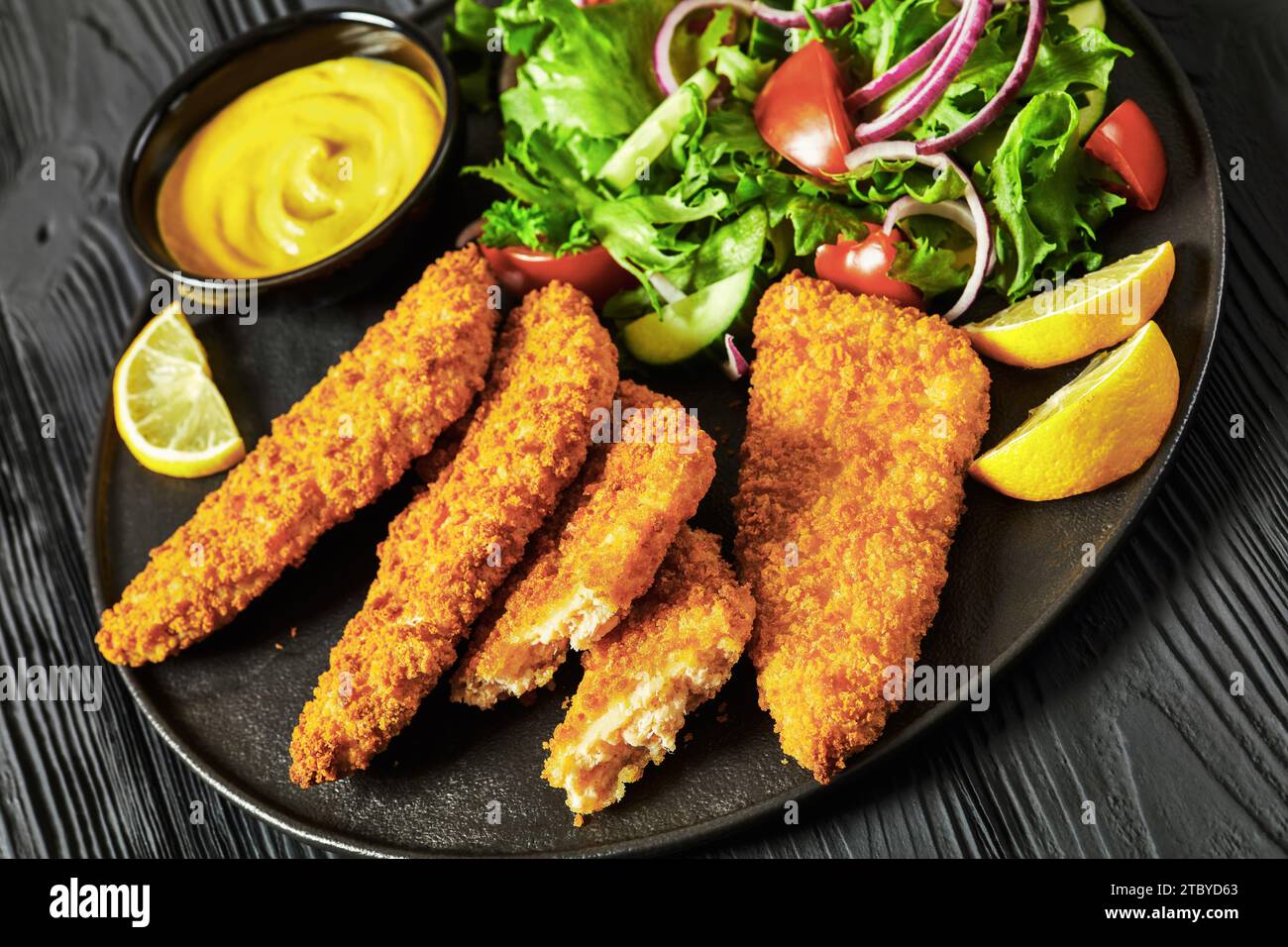 close-up of breaded fish fillet baked in oven served with yellow mustard, tortilla chips and fresh salad of lettuce, tomatoes, cucumber on black plate Stock Photo