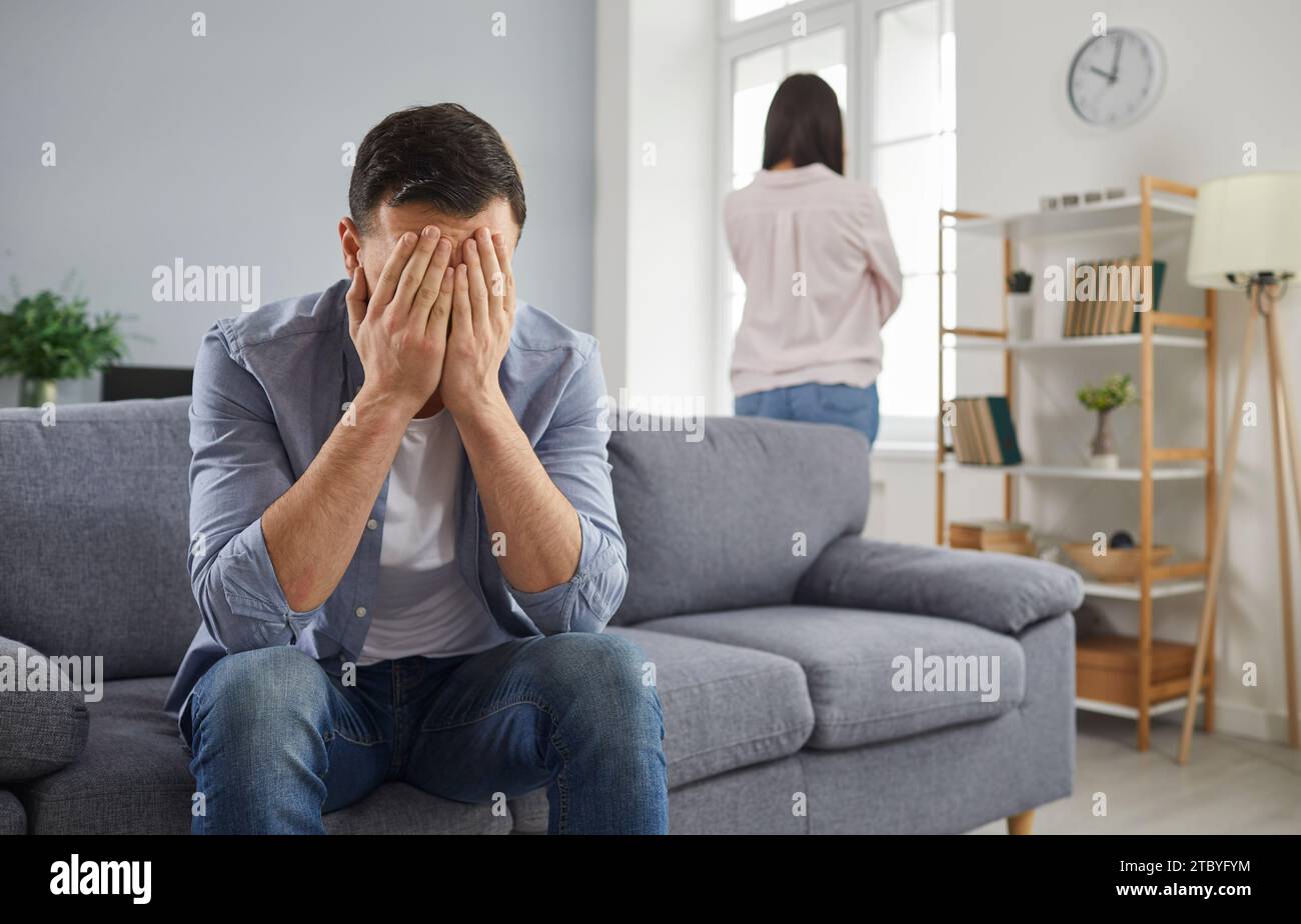 Upset young man covering his face with hands sitting on sofa with a woman in background at home. Stock Photo