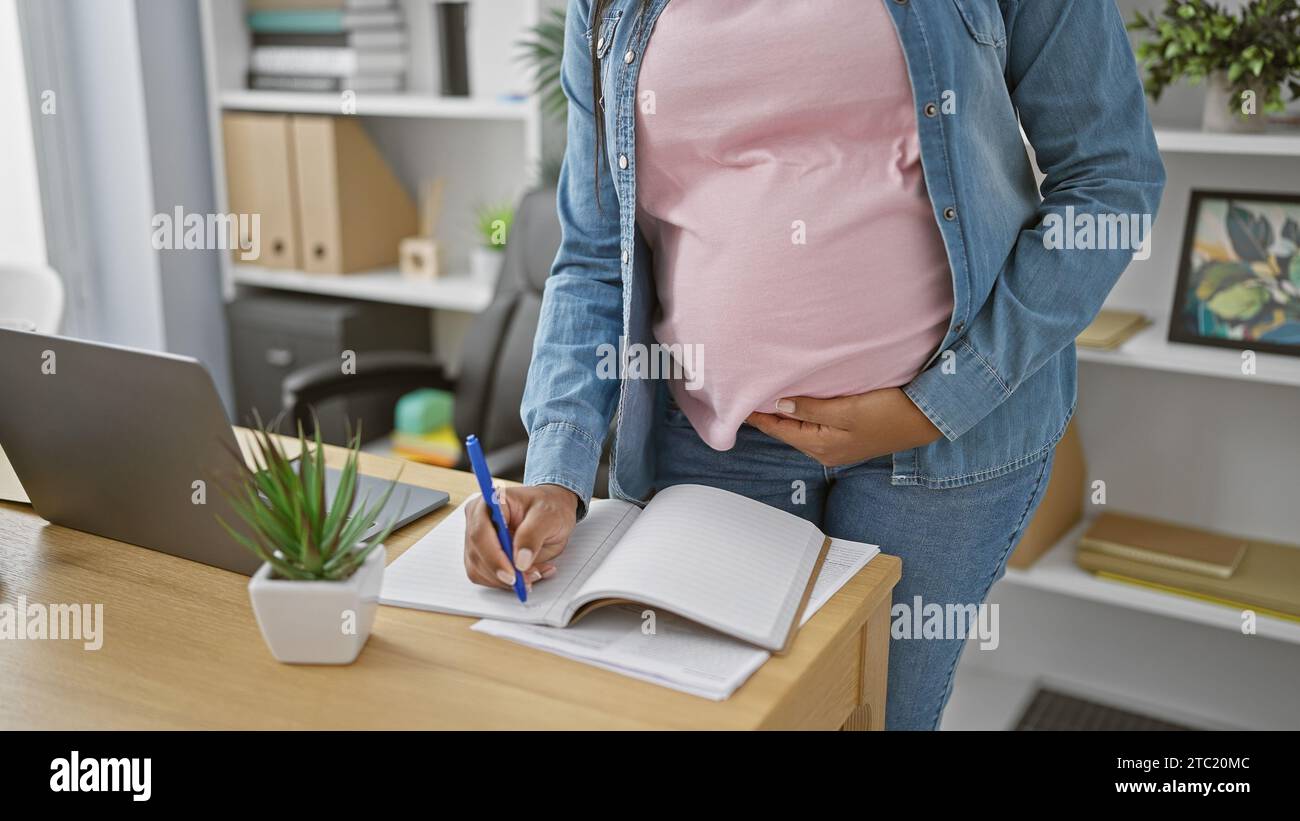 Expecting joy, young, hispanic working-mum-to-be massages belly while taking notes at office Stock Photo