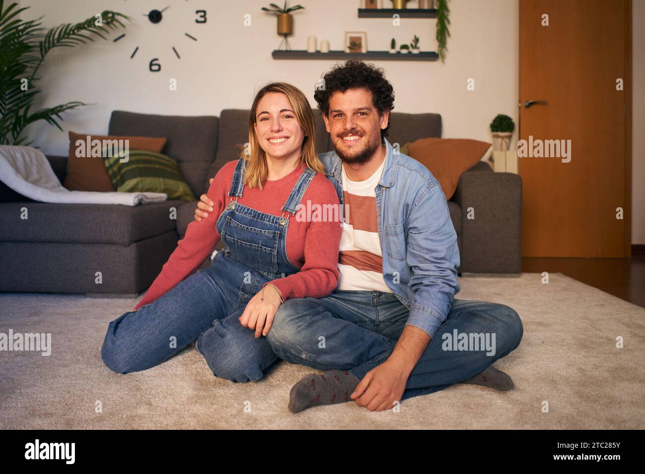 Two Caucasian joyful young adult in love looking at smiling camera sitting on carpet in living room. Stock Photo
