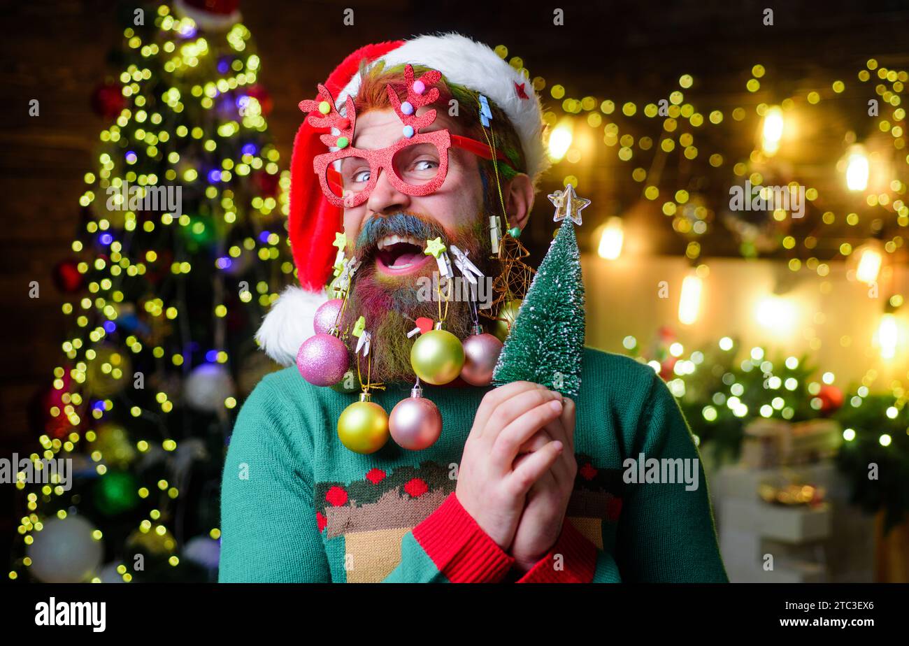 New year guy in Santa hat and party glasses with decoration balls in beard holds small Christmas tree. Christmas or New Year celebration. Christmas Stock Photo