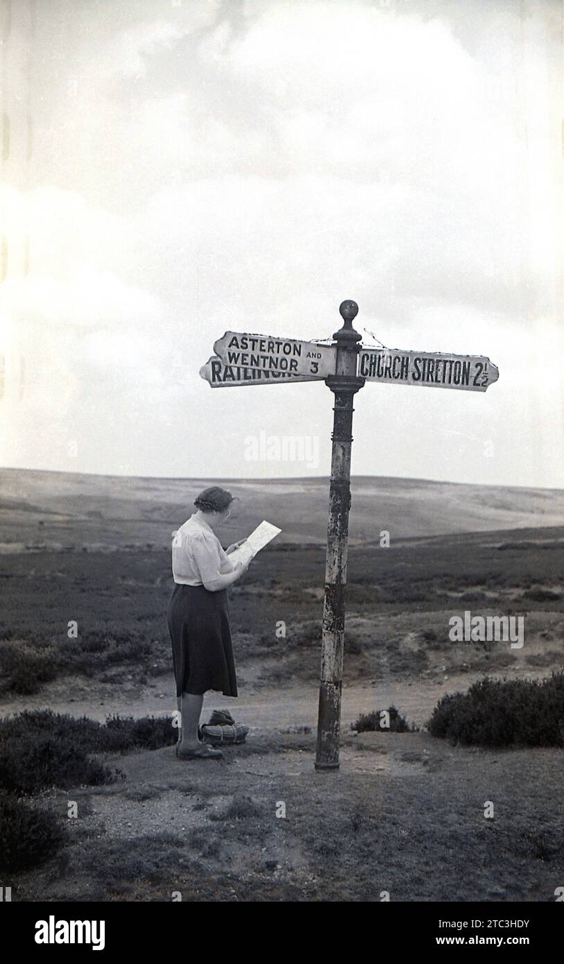 1950s, historical, a lady studying a map standing by a roadsign in the Shropshire Hills, England, UK. One directional arrow says Church Stretton, a market town in Shropshire, often referred to 'Little Switzerland' in the Victorian times, because of its landscape and picturesque setting in the Shropshire Hills. Stock Photo