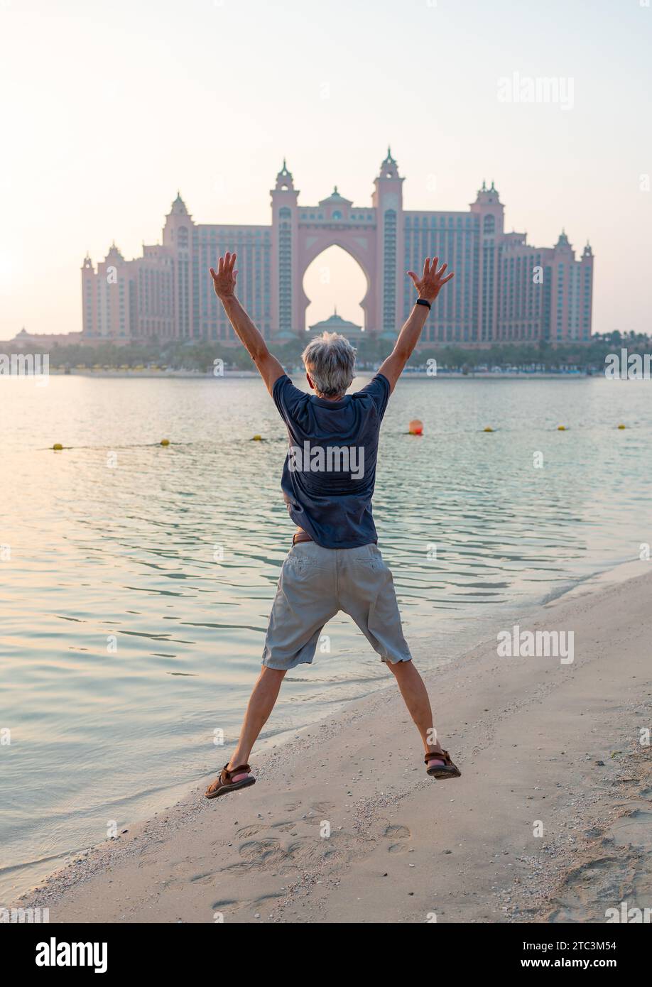 Senior caucasian man jumping high at Palm Jumeirah in Dubai, captured against the stunning backdrop of a sunset on the public beach Stock Photo