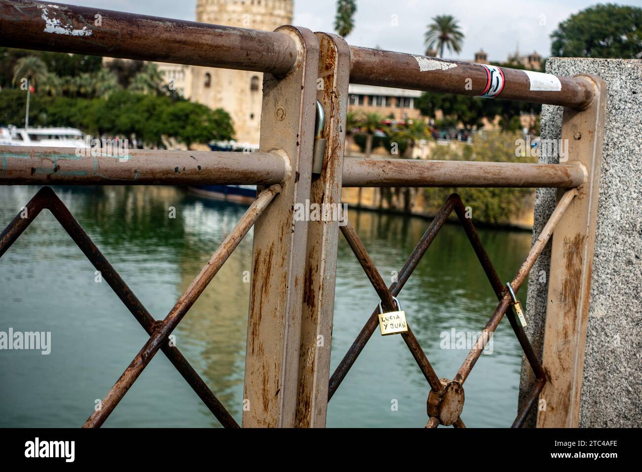 A close-up view of love locks attached to a rusty railing with Torre del Oro, Seville, Spain,  in the background. Stock Photo