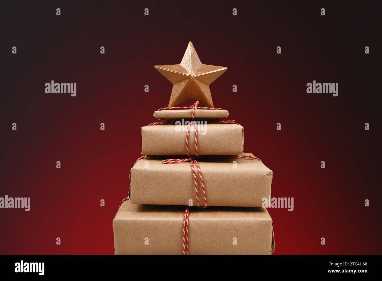 A stack of plain paper wrapped christmas presents on tied with twine and a gold star on top, against a light to dark red spot background. Stock Photo