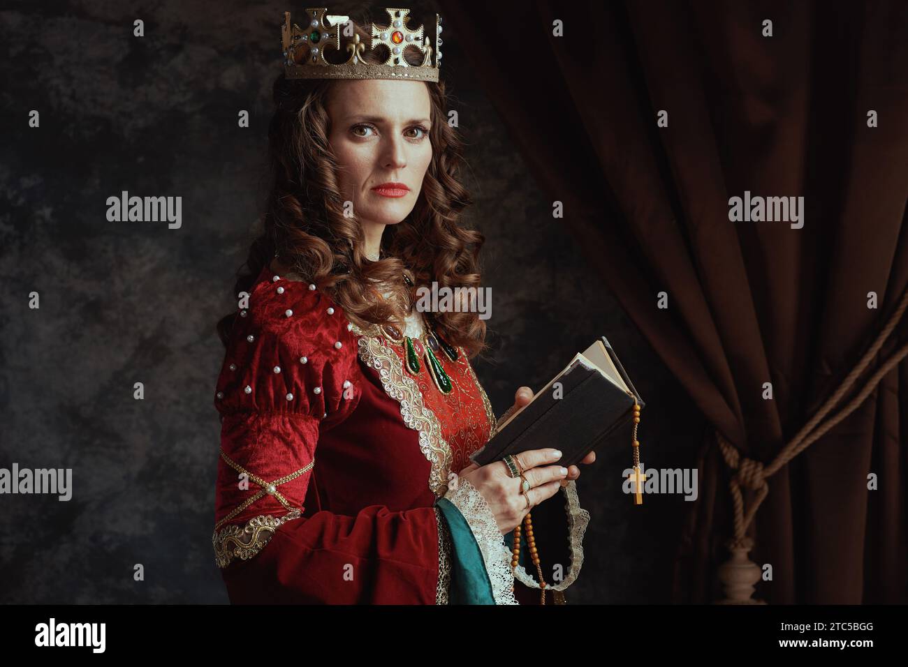 medieval queen in red dress with book, rosary and crown on dark gray ...