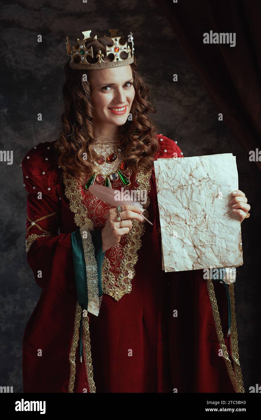 smiling medieval queen in red dress with parchment and crown on dark gray background. Stock Photo