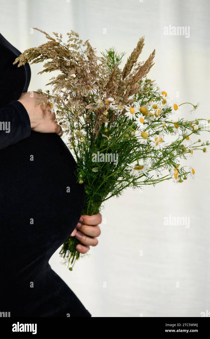 A pregnant woman is hugging her belly against a white background and wearing a black bodycon dress and holding a bouquet of wildflowers in one hand. Stock Photo