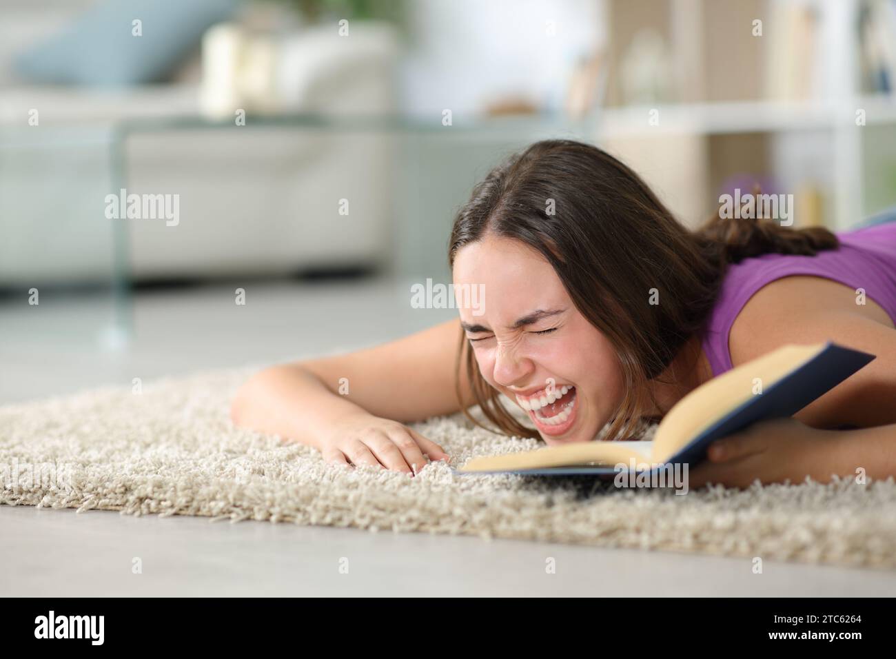 Happy woman lying on the floor laughing hilariously reading a book at home Stock Photo