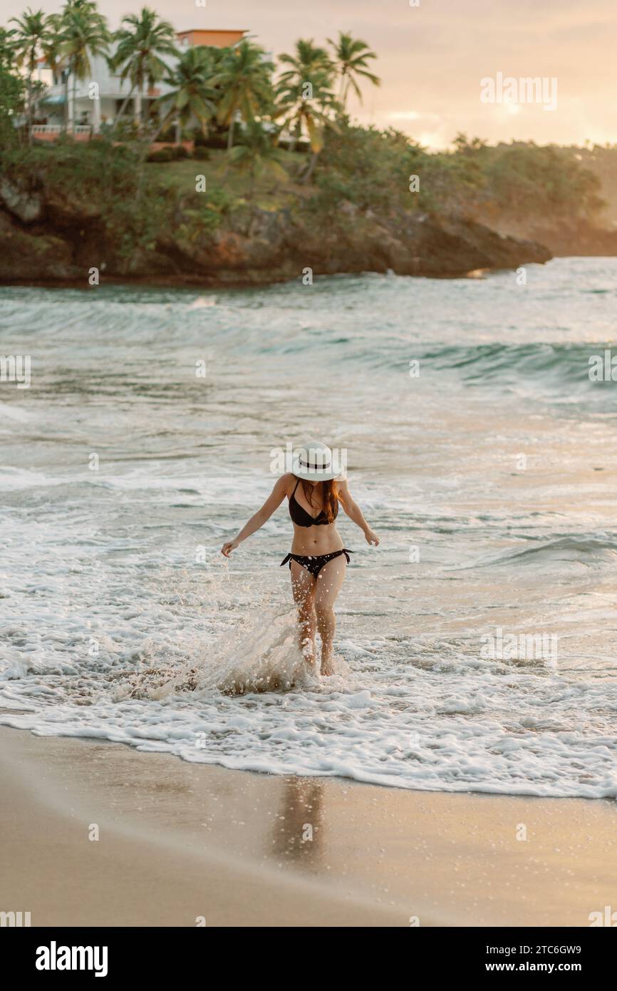Joyful woman in black bikini and hat plays in surf at tropical sunset. Stock Photo