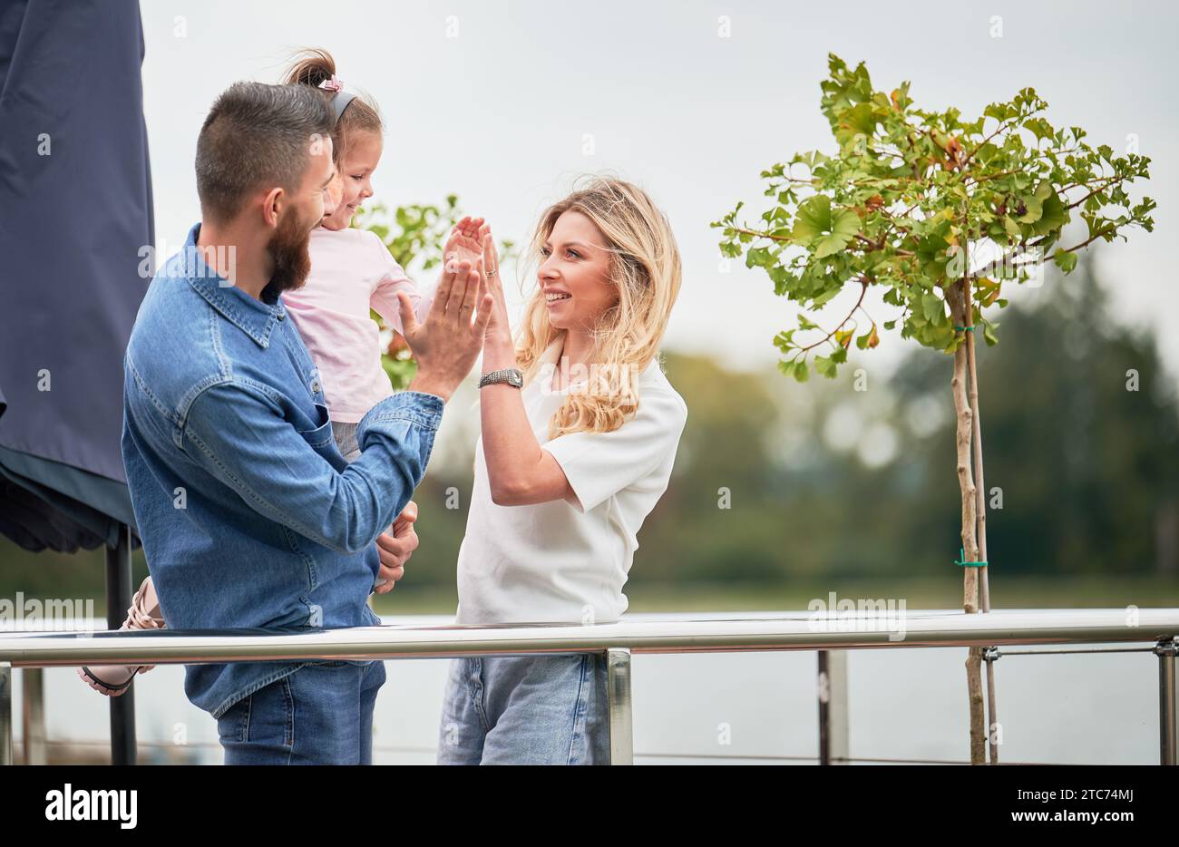 Cheerful woman, man and little girl standing near green tree and slapping hands giving high five outdoors. Happy man holding daughter and smiling while celebrating success with wife and child. Stock Photo