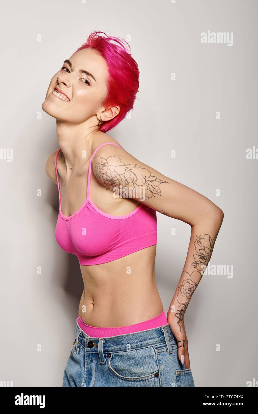 tattooed and expressive woman with pink hair posing with hands in pockets of jeans on grey Stock Photo