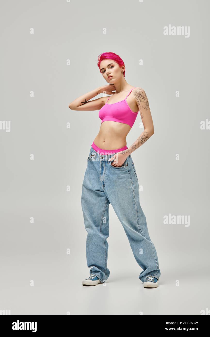 appealing young woman with pink hair and tattoos in stylish crop top posing on grey backdrop Stock Photo