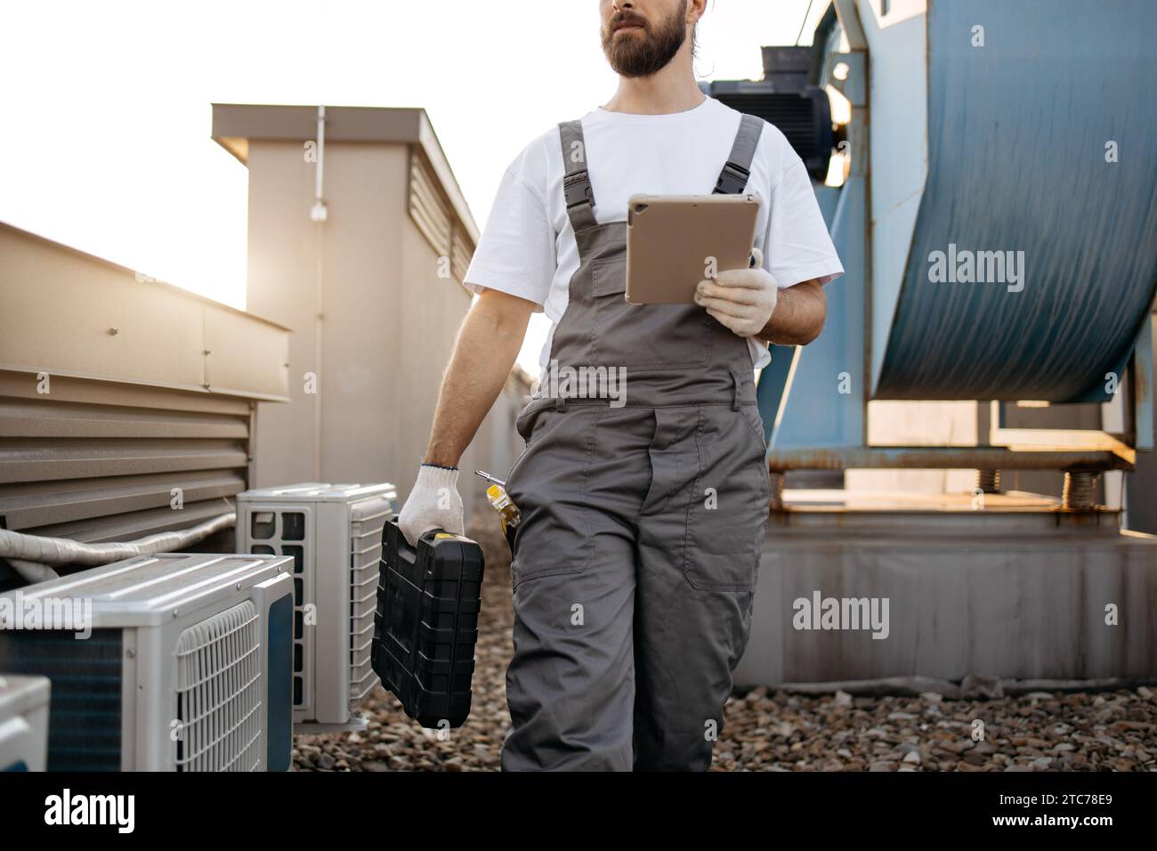 Close up of bearded repairman dressed in gray overall walking and holding wireless tablet in hand while carrying black case with tools on factory terr Stock Photo