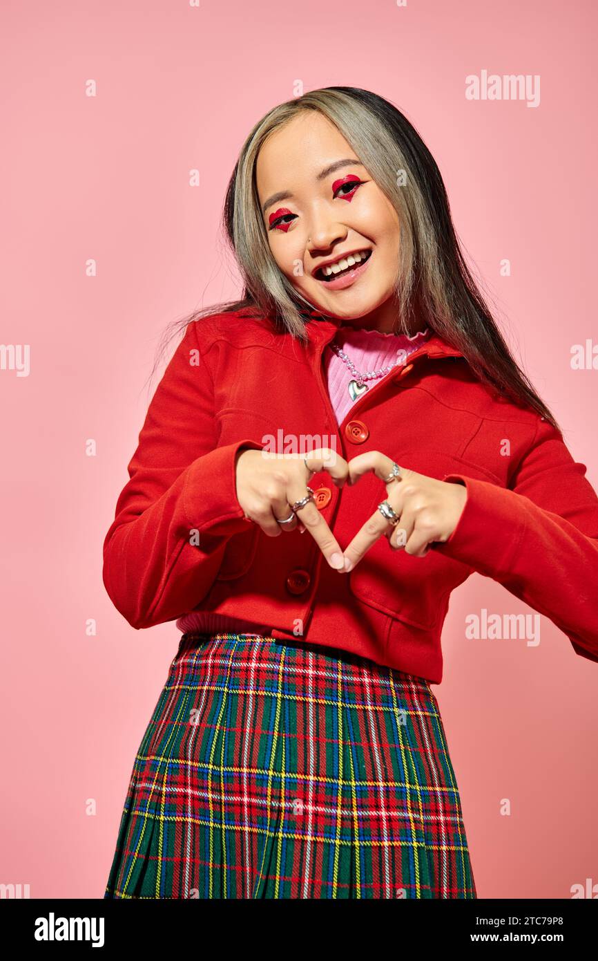 Valentines day concept, happy asian girl in red jacket showing heart with fingers on pink backdrop Stock Photo