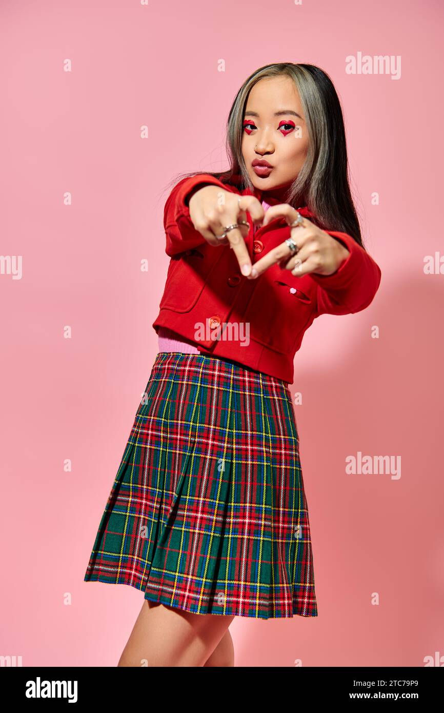 Valentines day concept, young asian girl in red jacket showing heart with fingers on pink backdrop Stock Photo