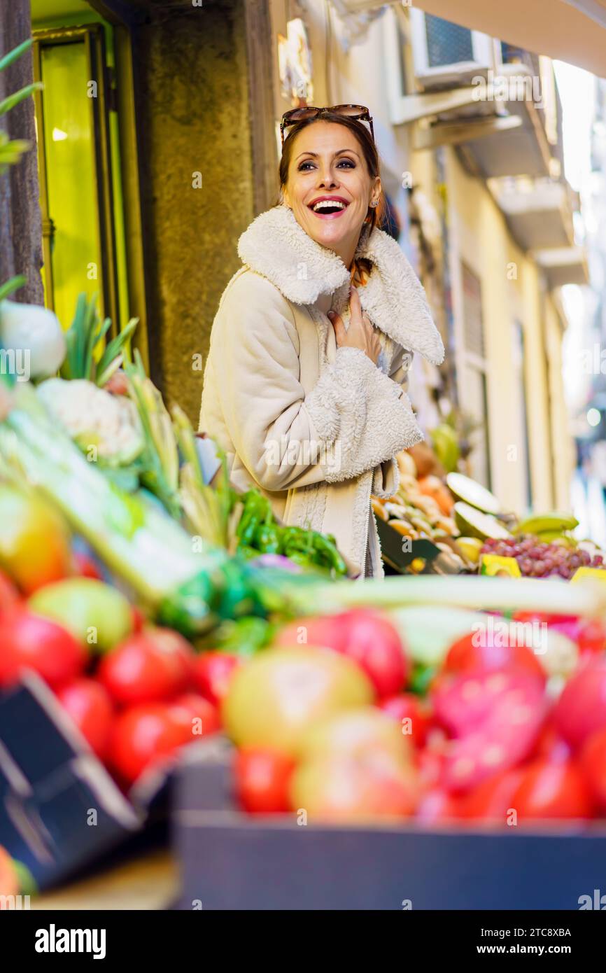 Gleeful young woman in cozy coat standing by fruits in shop Stock Photo