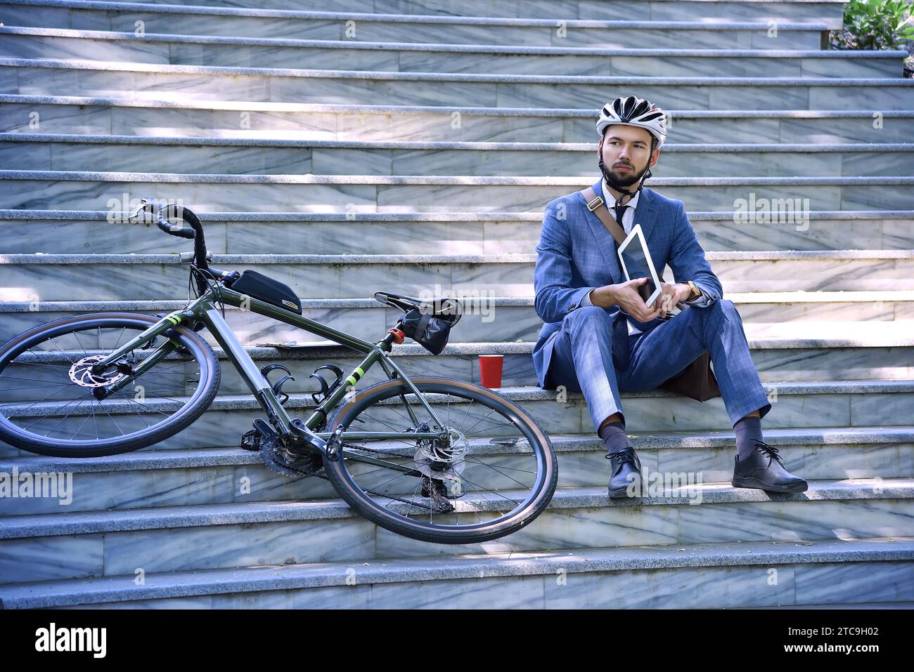 Businessman sitting on stairs with bicycle, helmet and using digital tablet. Business and alternative transport concept Stock Photo