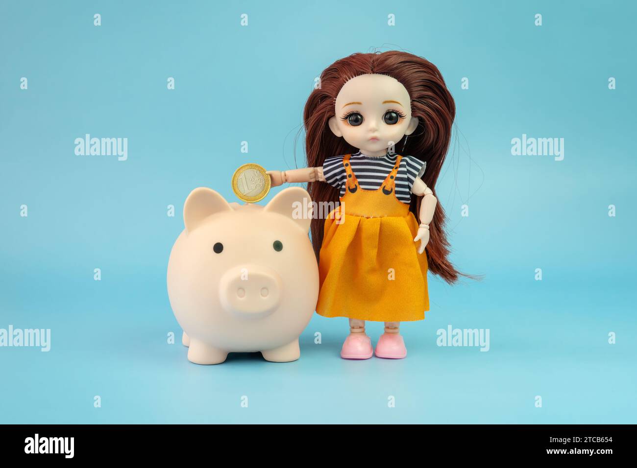 A small child doll with a coin in her hands on a blue background. The doll puts a coin in the piggy bank. Stock Photo