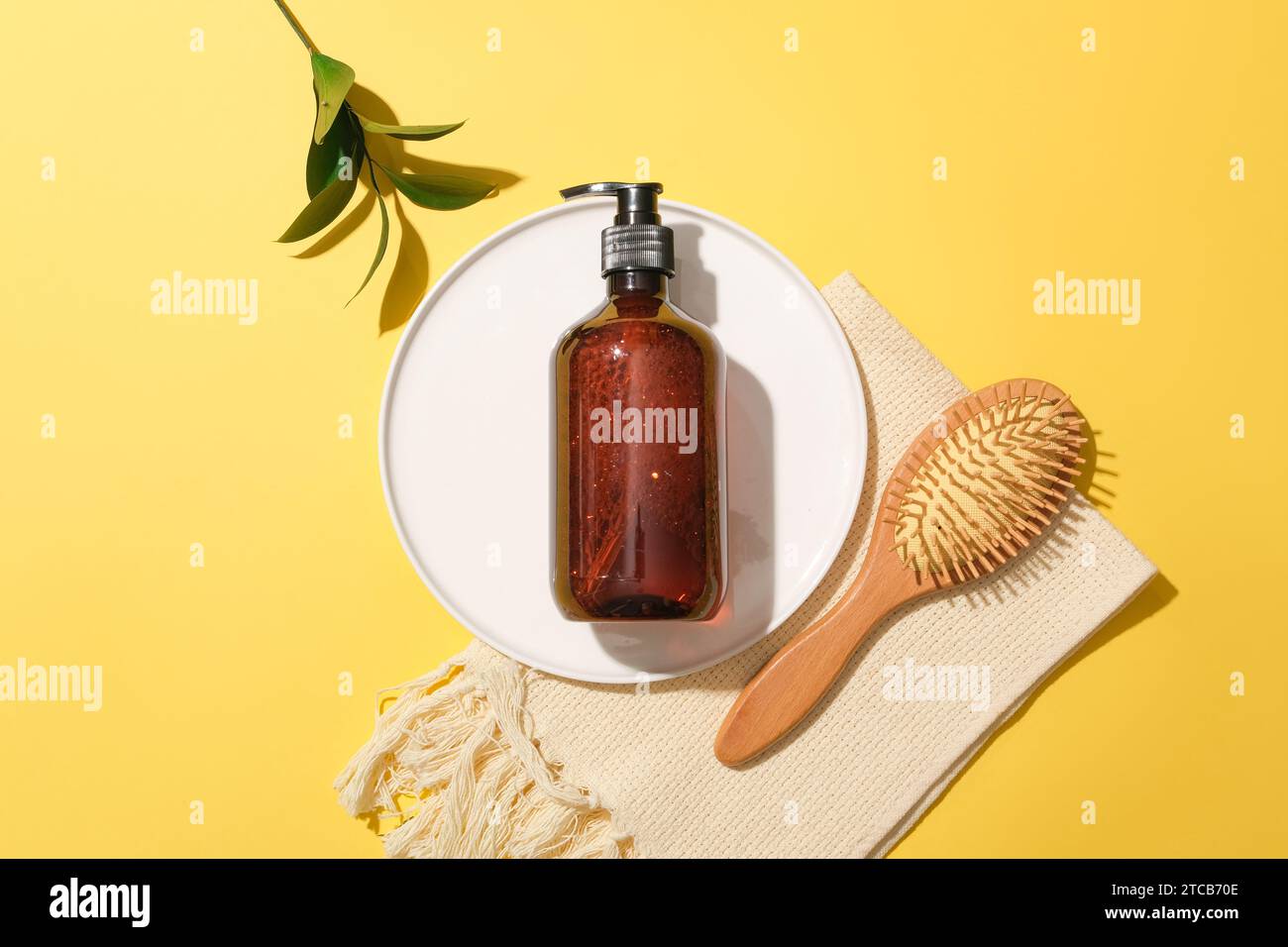 View from above of a brown shampoo bottle on a white ceramic plate, a detangling comb, green leaves and a towel on a yellow background. Cosmetic mocku Stock Photo