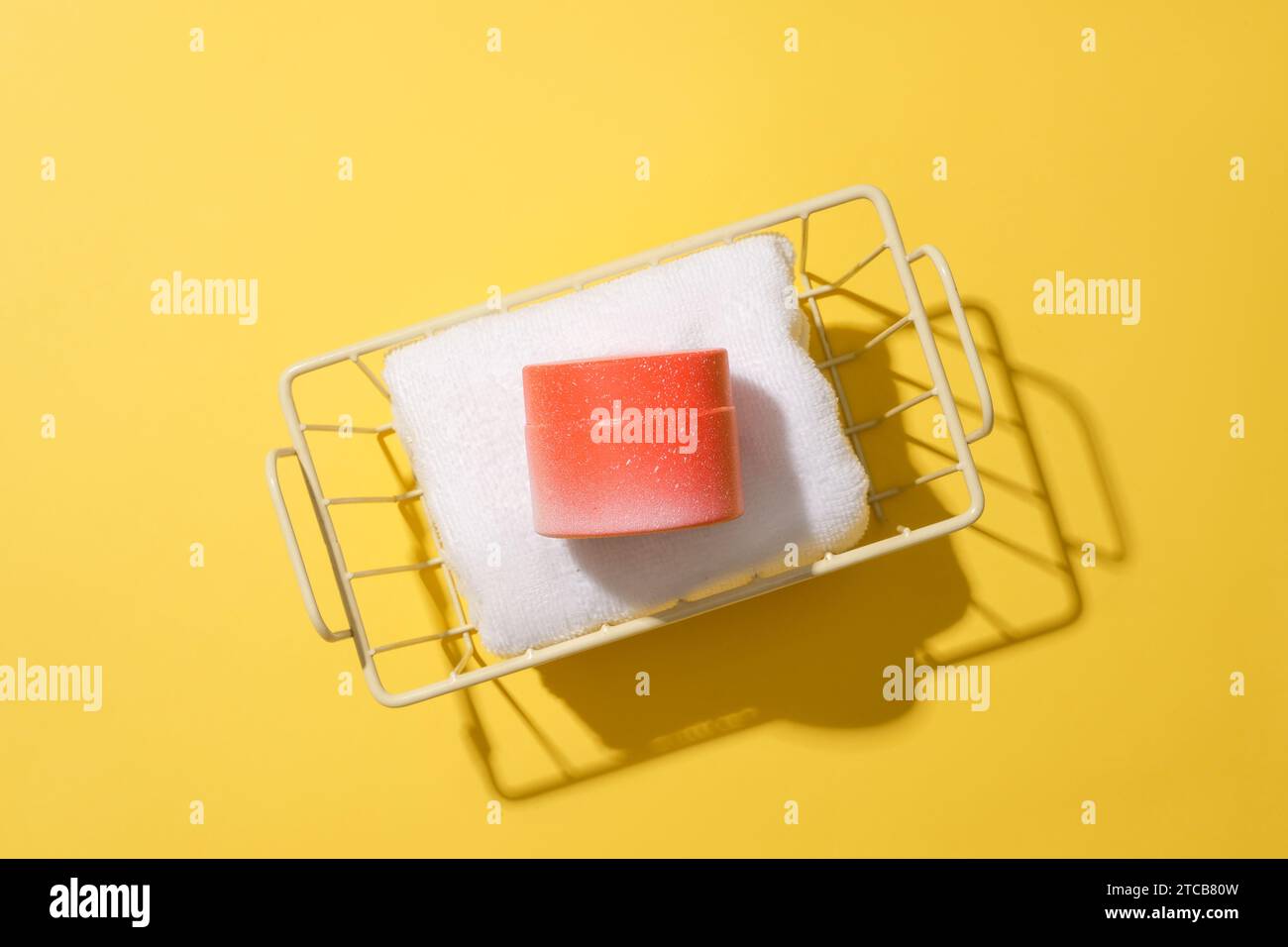 Close-up of a cosmetic jar lying on a towel rolled up in an iron mesh basket. Bright yellow background. Cosmetic mockup for advertising. Blank label f Stock Photo