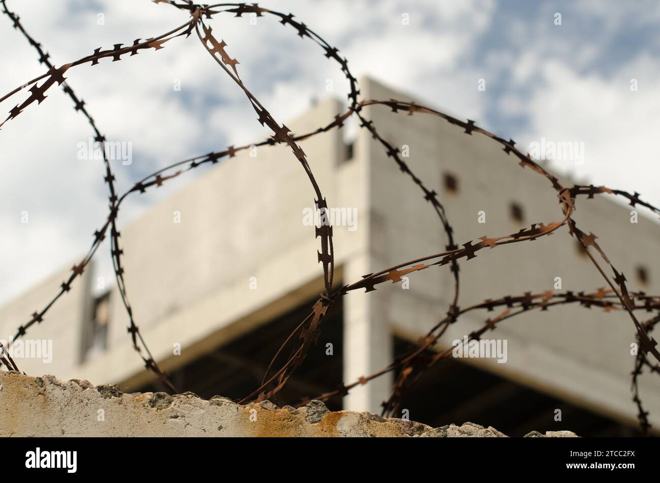 Old rusty barbed wire fence against the backdrop of an abandoned building and the blue sky Stock Photo