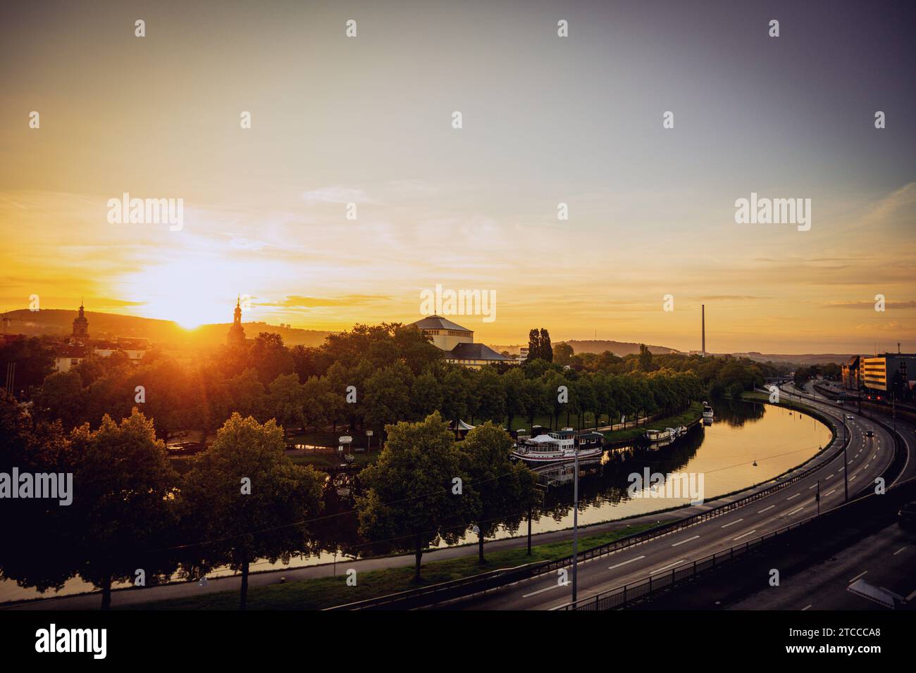 A view of a bustling urban landscape, featuring several cars driving down a road which leads to a stunning sunset over the water Stock Photo