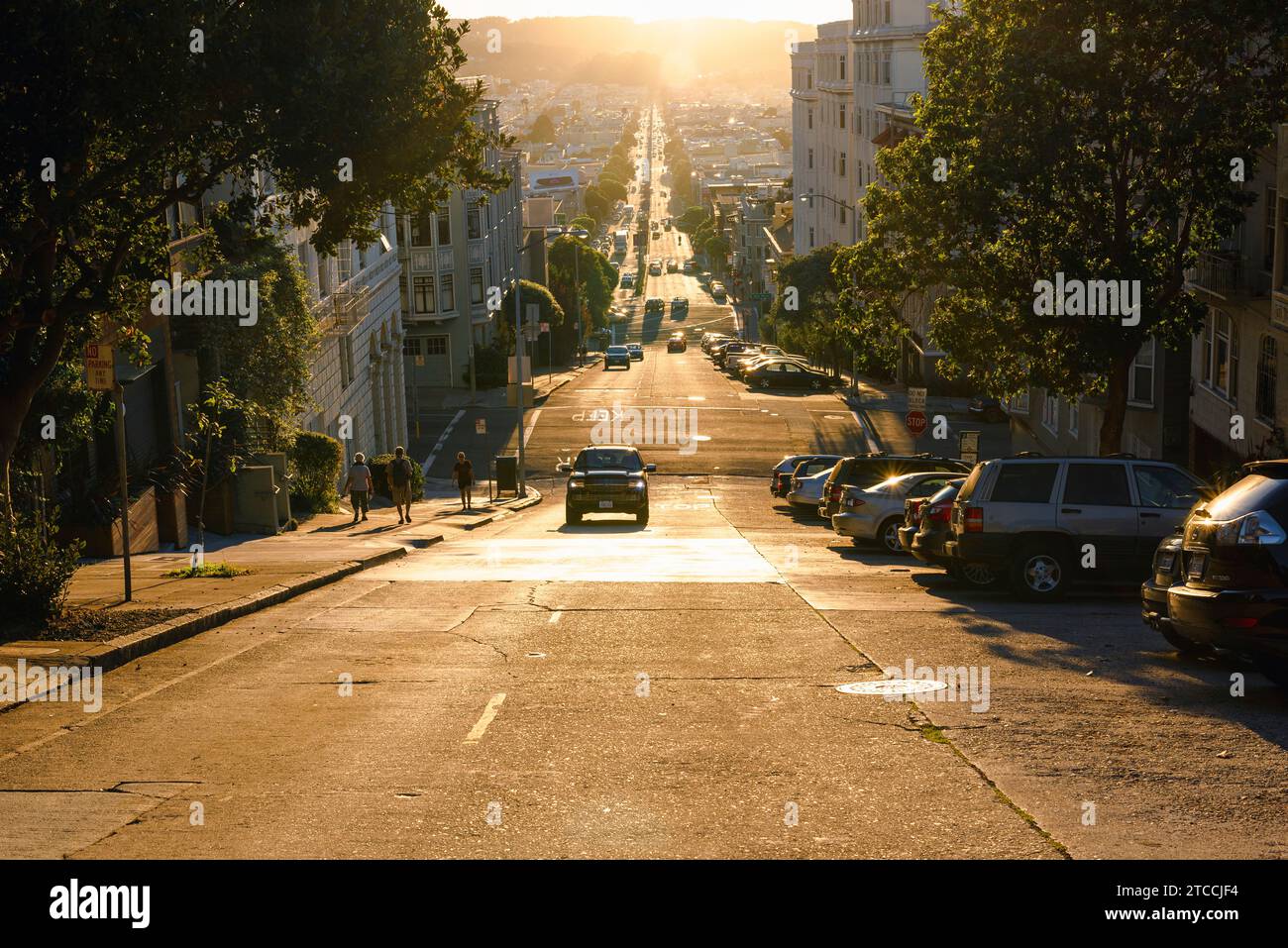 A car climbs one of the steep hills in San Francisco as the sun sets over the city in the background. Stock Photo