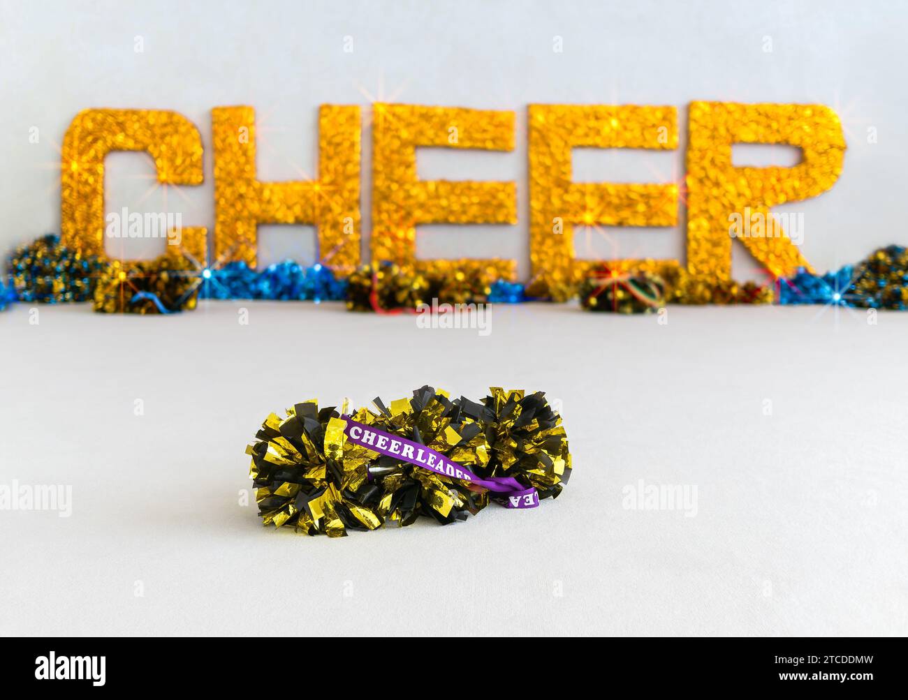 Gold and black pom poms with 'Cheerleader' written on a purple ribbon and large gold sparkly 'CHEER' letters in the background Stock Photo