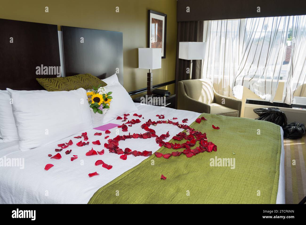 A white bed with rose petals arranged in a heart shape on the bedspread. Stock Photo