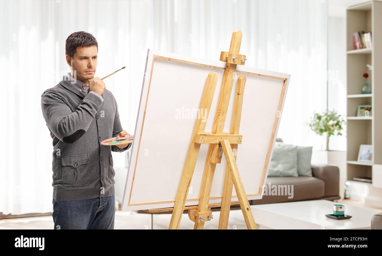 Pensive man in front of a painting canvas with a brush and palette at home in a living room Stock Photo