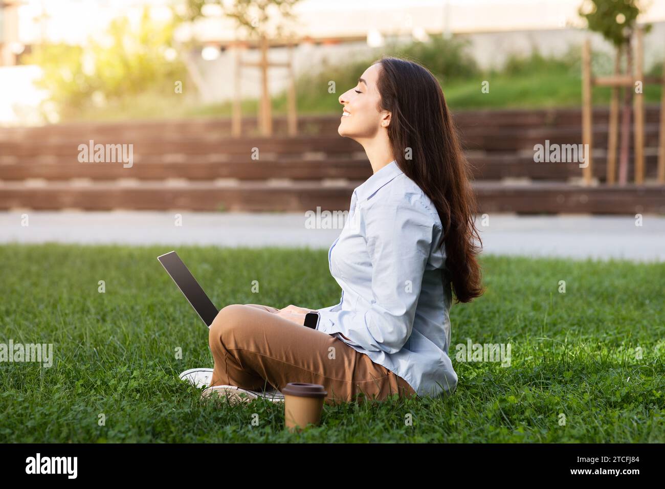 Positive serene professional european young woman sits with laptop in grassy outdoor area Stock Photo