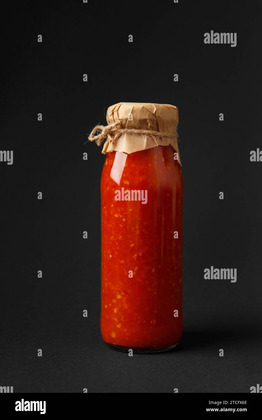 Hot sauce made from chili peppers, paprika, tomatoes and garlic in a glass jar, isolated on a black background. Caucasian sauce, adjika. Stock Photo
