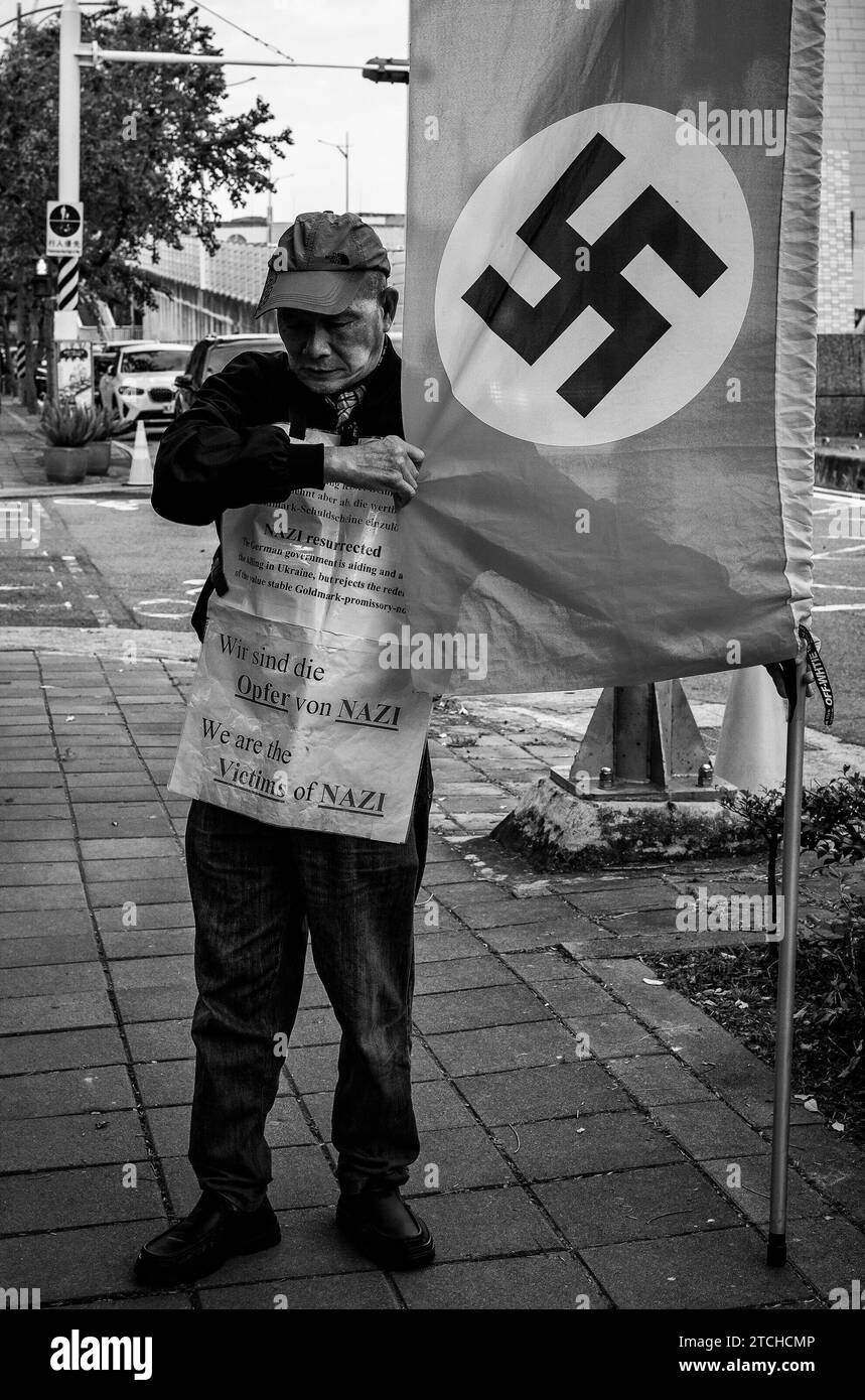 Taipei. 13th Dec, 2023. A protester stands with a flag emblazoned with the Nazi symbol in front of the European School in Taipei, Taiwan, on 13/12/2023, demanding the German government honor the bonds issued to Japan as reparations for World War I. The Old German Mark Association, comprised of descendants of Taiwanese who purchased these bonds during Japanese colonial rule, asserts that despite the German and Japanese governments' refusal to redeem the bonds, their financial and historical value remains by Wiktor Dabkowski Credit: dpa/Alamy Live News Stock Photo