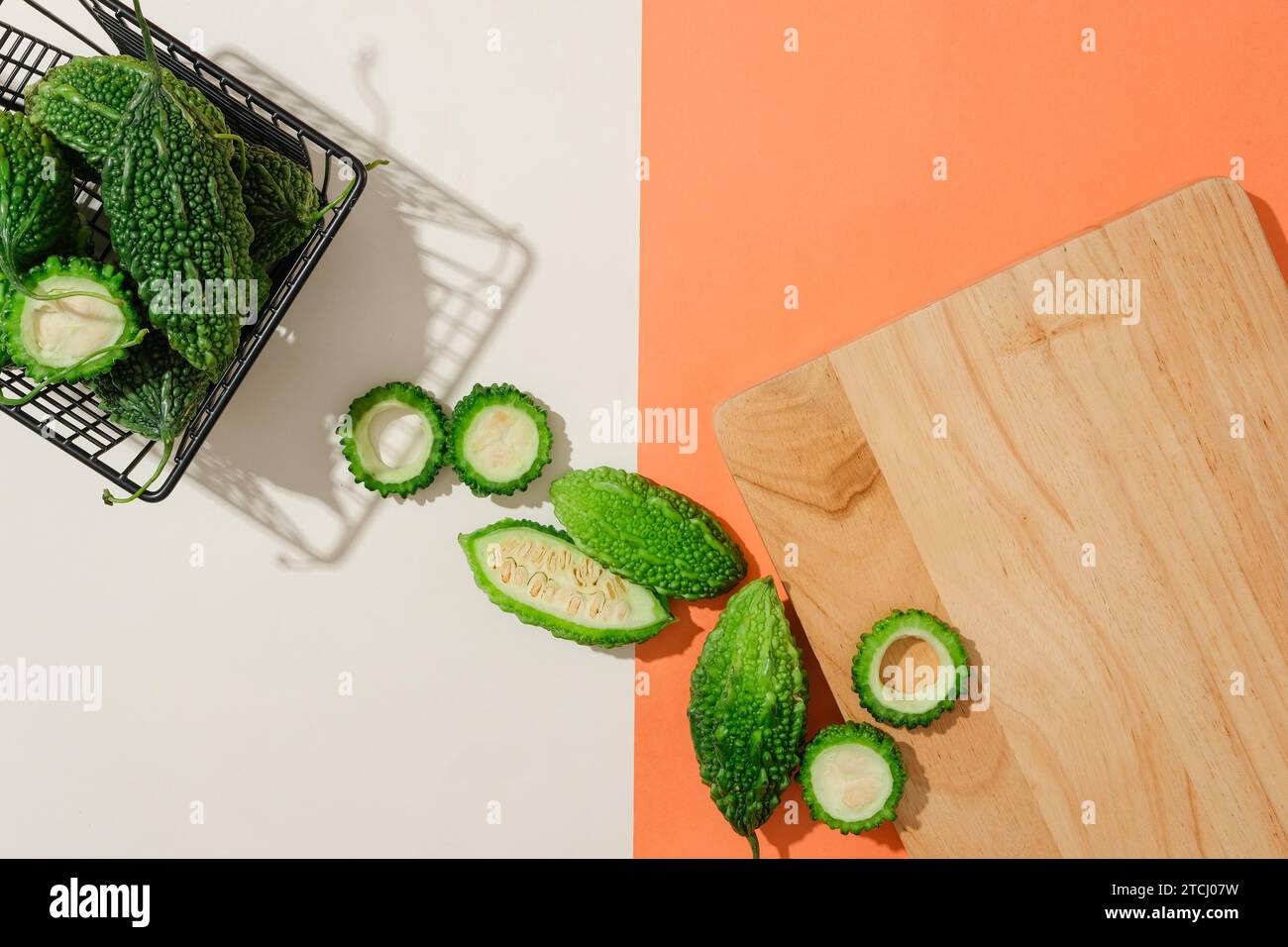 The background is divided into two halves with two colors red and white. Bitter melon is stored in an iron mesh basket and a wooden cutting board is d Stock Photo