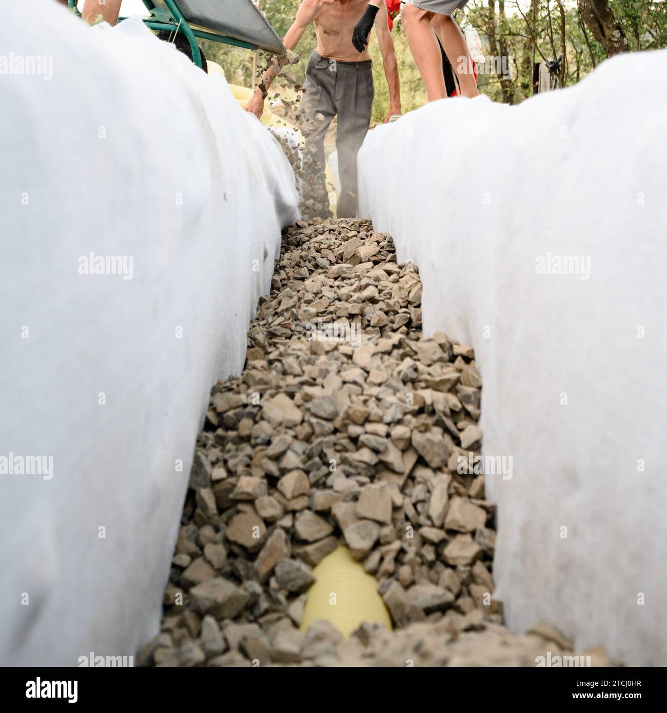 A man pours rubble crushed stone from a wheelbarrow into a trench. Drainage works for drainage of ground water around the field. Stock Photo