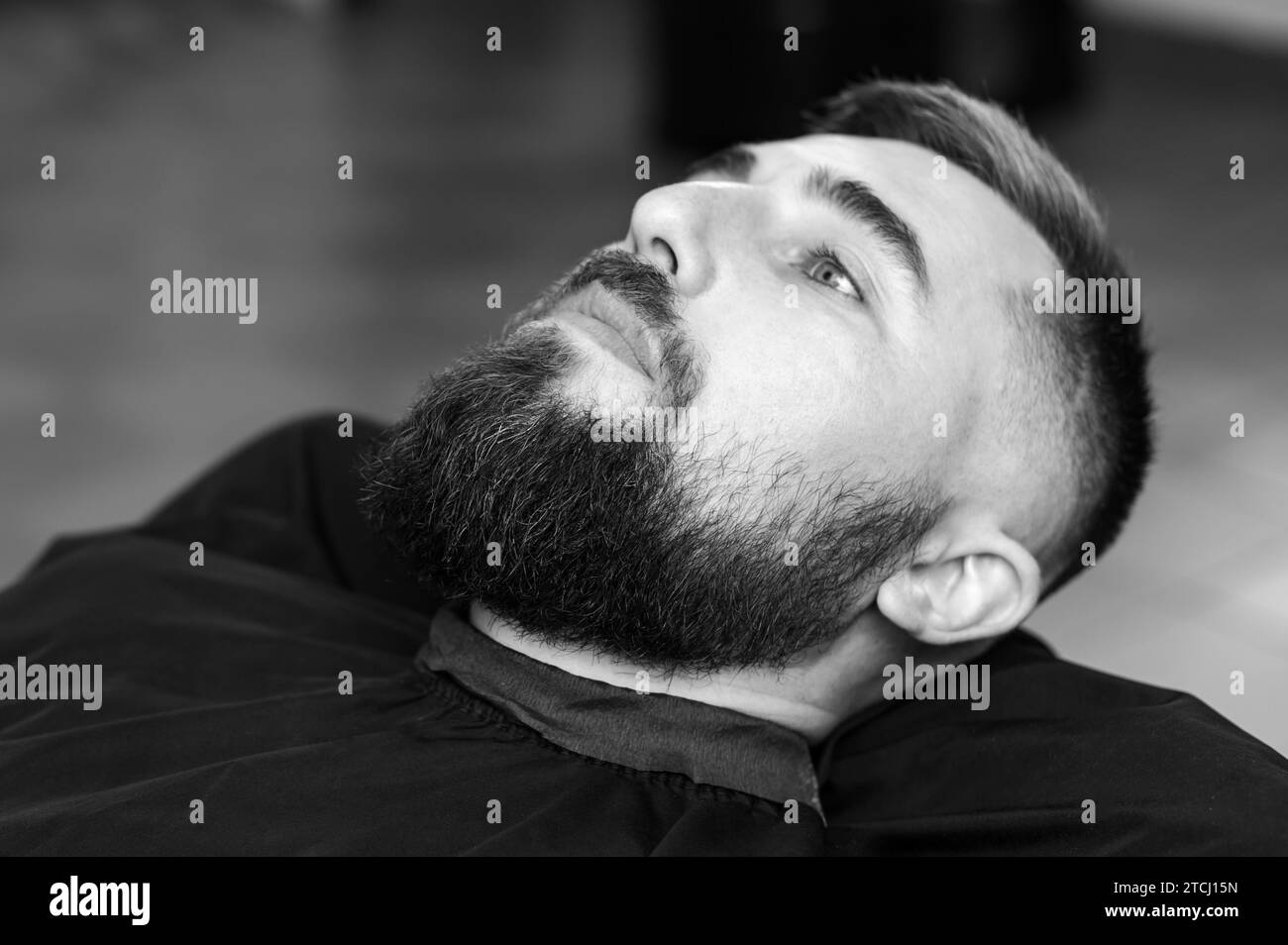 A young man with a beard in a black cape sits on a chair in a barbershop. The client is waiting for a haircut and shaving of the beard. Stock Photo