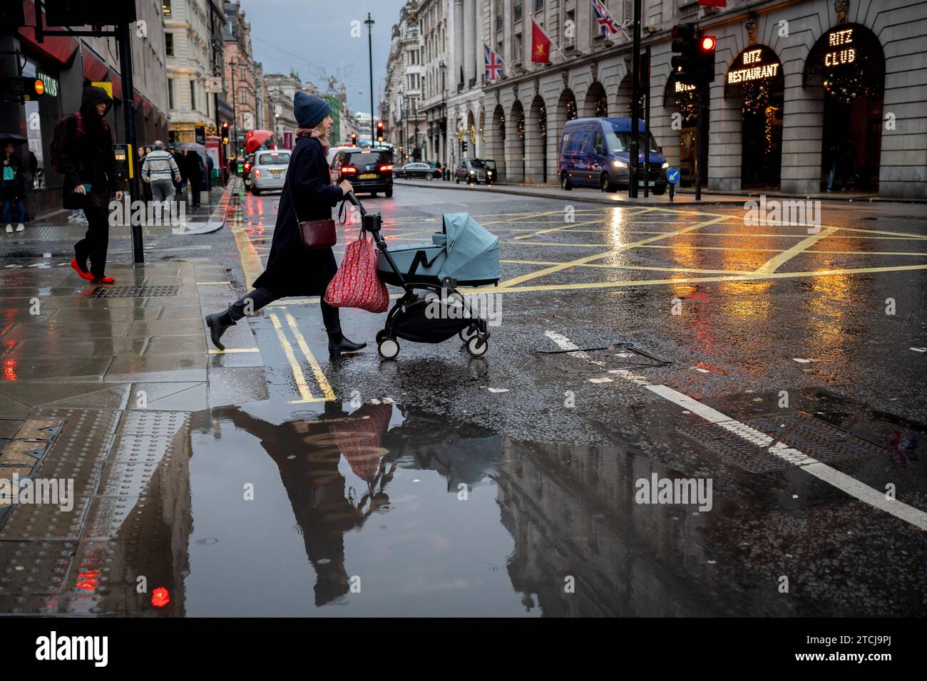 A woman pushes a child in its pushchair and crosses next to a puddle to cross Piccadilly on a rainy afternoon  in Westminster, on 12th December 2023, in London, England. Stock Photo
