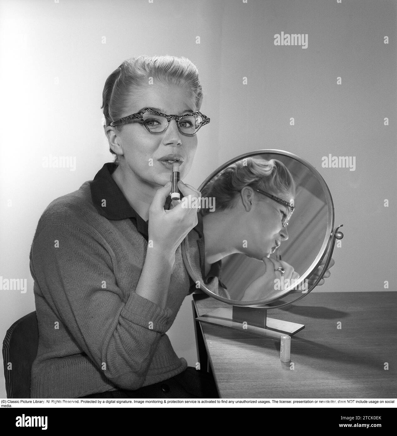 In the 1950s. A young woman with a mirror putting lipstick on. She has the glasses and bows typical of the 1950s. 1958. Kristoffersson ref CB99-11 Stock Photo