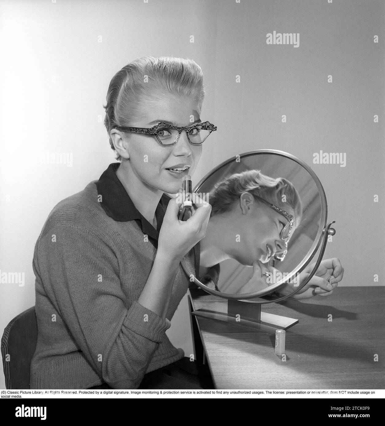 In the 1950s. A young woman with a mirror putting lipstick on. She has the glasses and bows typical of the 1950s. 1958. Kristoffersson ref CB99-12 Stock Photo