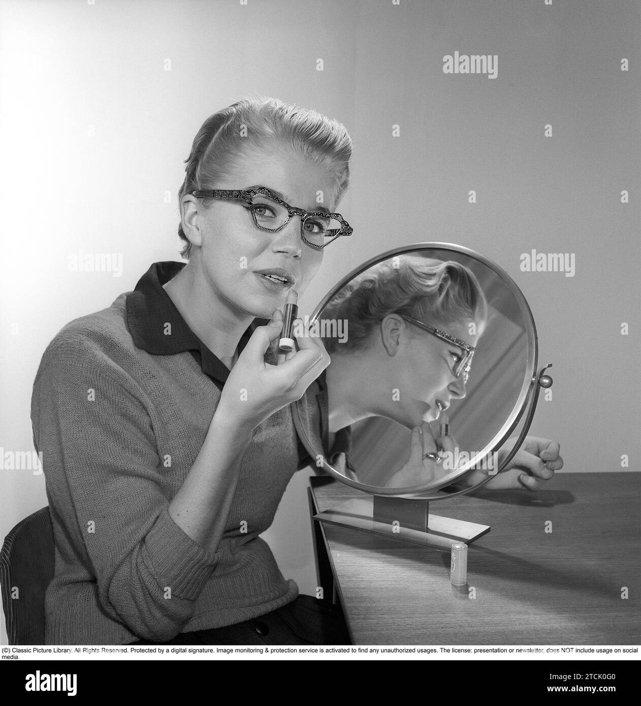 In the 1950s. A young woman with a mirror putting lipstick on. She has the glasses and bows typical of the 1950s. 1958. Kristoffersson ref CB99-12 Stock Photo