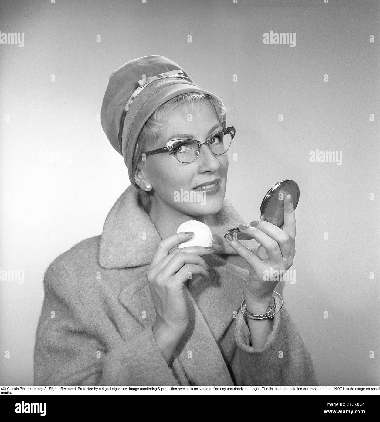1950s makeup. A woman is looking at herself in her pocket mirror and improves on her makeup. She is wearing a fashionable hat, typical 1950s glasses and a coat. 1958 Kristoffersson ref CB96-5 Stock Photo