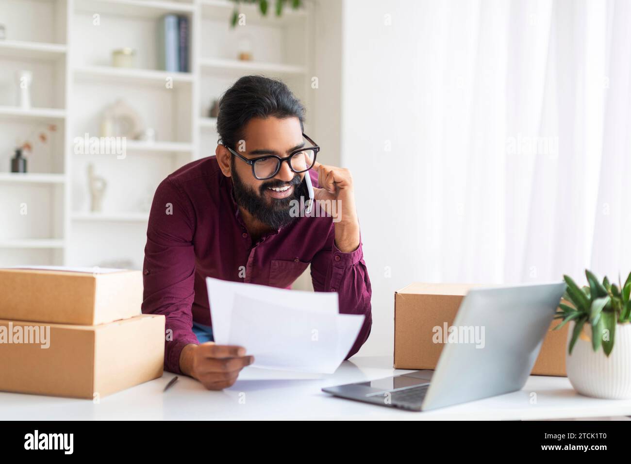 Smiling Indian freelancer verifying shipping documents while on phone call Stock Photo