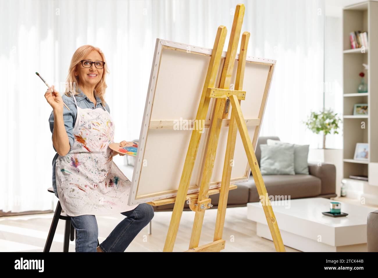 Woman sitting on a chair holding a painting brush and color palette in front of a canvas at home Stock Photo