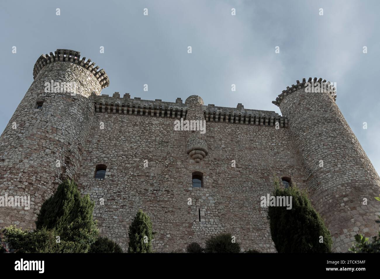 stone castle with two towers and a wall with battlements stands against a gray, cloudy sky, surrounded by greenery in guadalajara spain Stock Photo