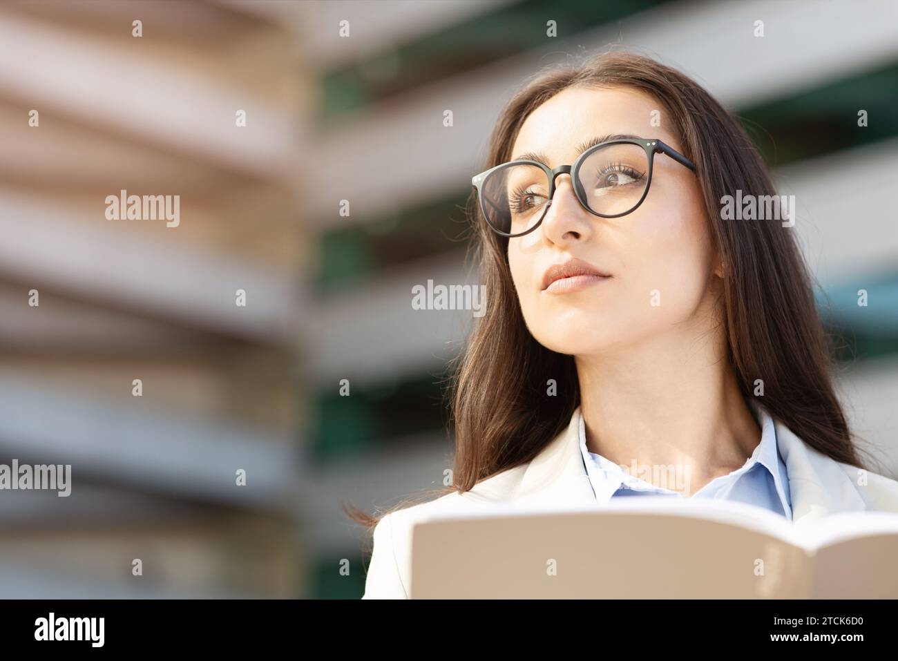 Thoughtful european millennial woman in glasses looks upward, holding open book, paper project Stock Photo