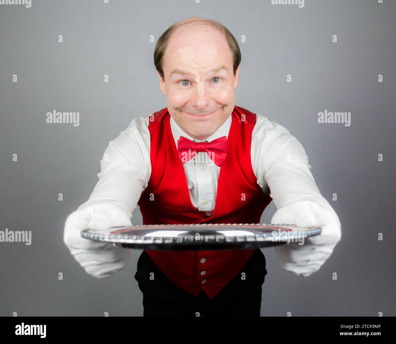 Portrait of Waiter or Servant in White Gloves and Red Vest or Waistcoat Smiling Kindly and Holding Silver Serving Tray. Professional Hospitality. Stock Photo
